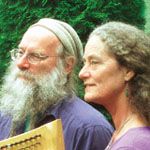 Two older adults with gray hair and glasses, the man holding a wooden harp, standing outdoors with green foliage in the background.
