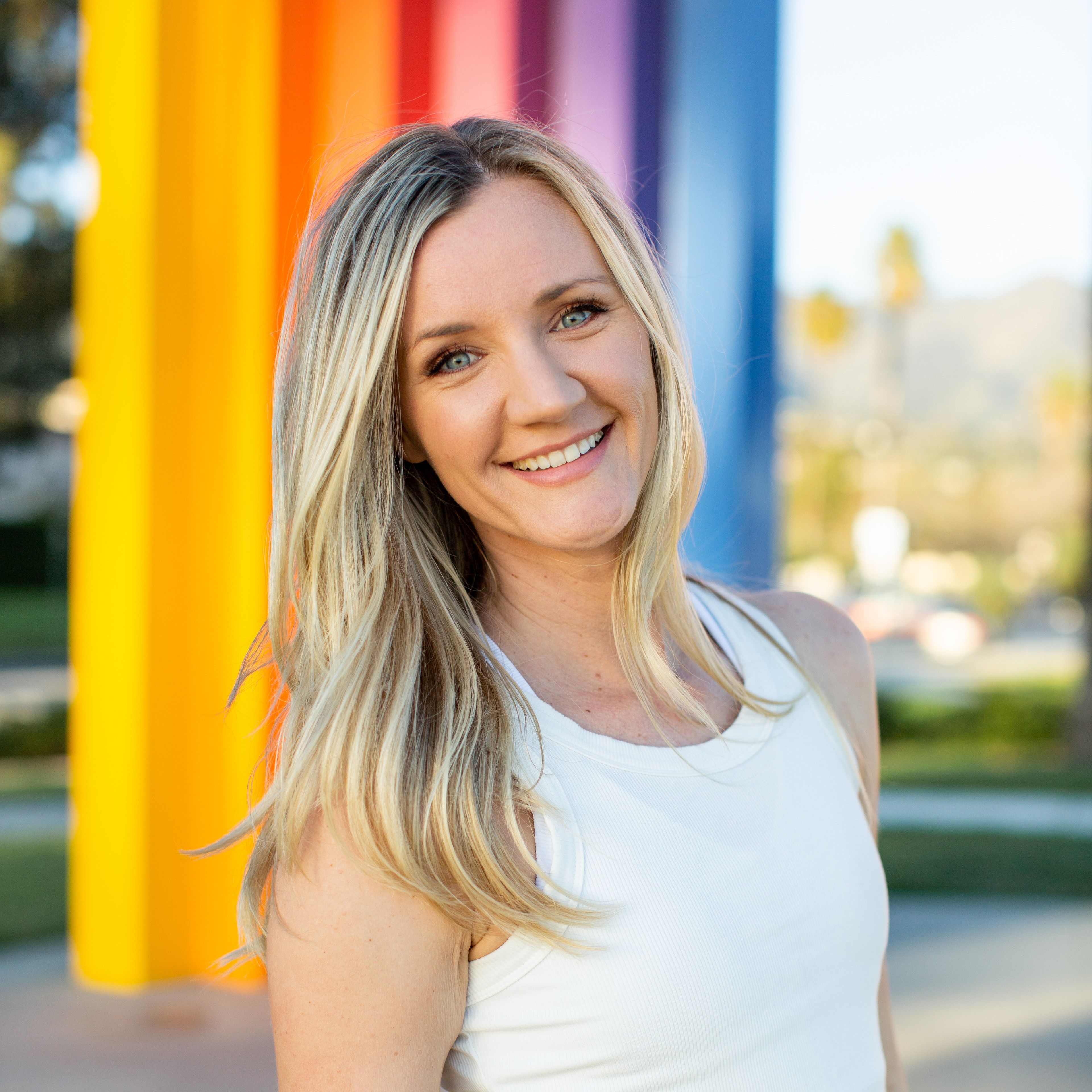 Smiling blonde woman with long hair wearing a white sleeveless top standing outdoors with colorful vertical pillars in the background.