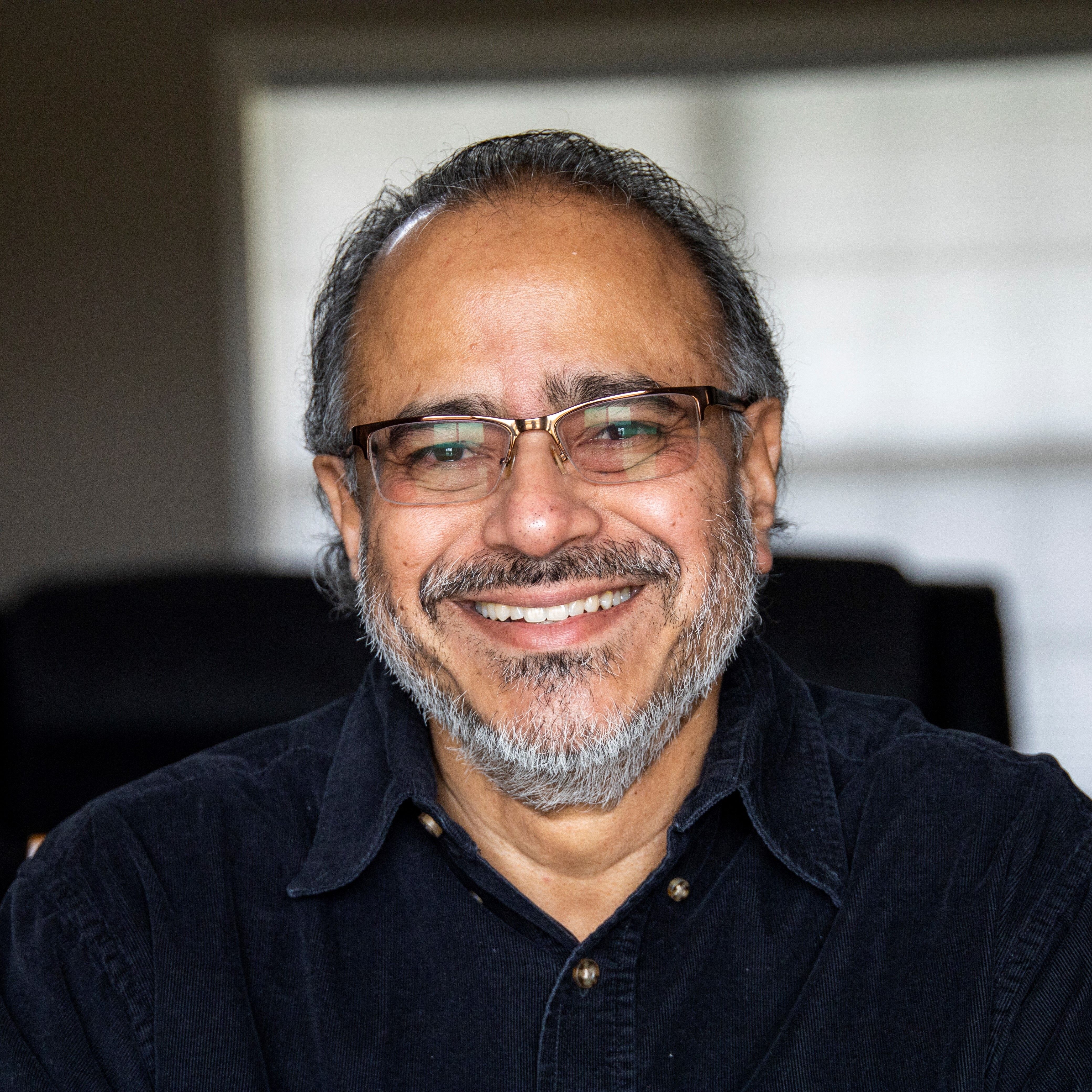Smiling middle-aged man with glasses, gray beard, and dark shirt against a blurred background.